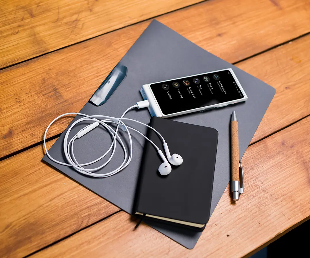 Vier Erwachsene, drei sichtbar und einer teilweise verdeckt, unterhalten sich im Freien am Wasser. Zwei Frauen, von denen eine lächelt und einen Hut trägt, zeigen der Gruppe etwas auf ihren Übersetzern, die sie in den Händen halten.