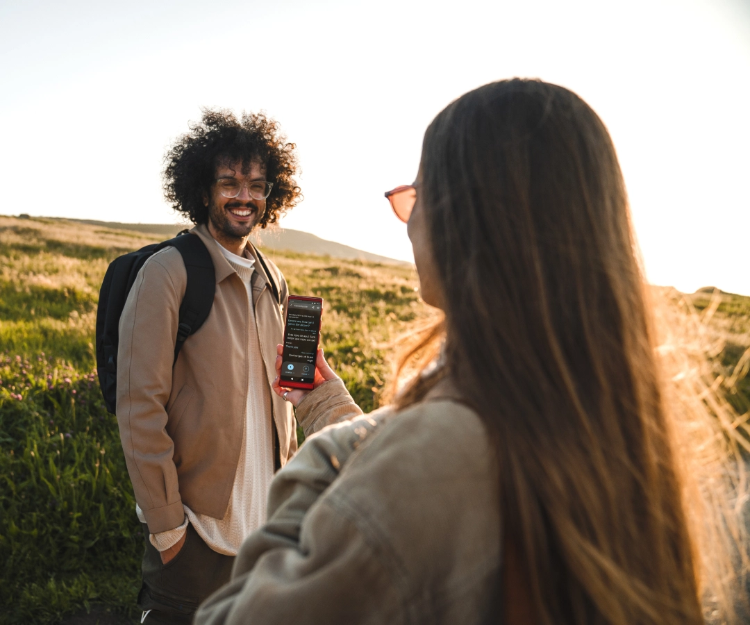 Ein Mann sitzt auf dem Fahrersitz eines Taxis und zeigt einer Frau auf dem Rücksitz etwas auf einem Übersetzer, den er in der Hand hält. Die Frau lächelt interessiert. Auf dem Dach des Wagens ist ein Taxischild zu sehen.