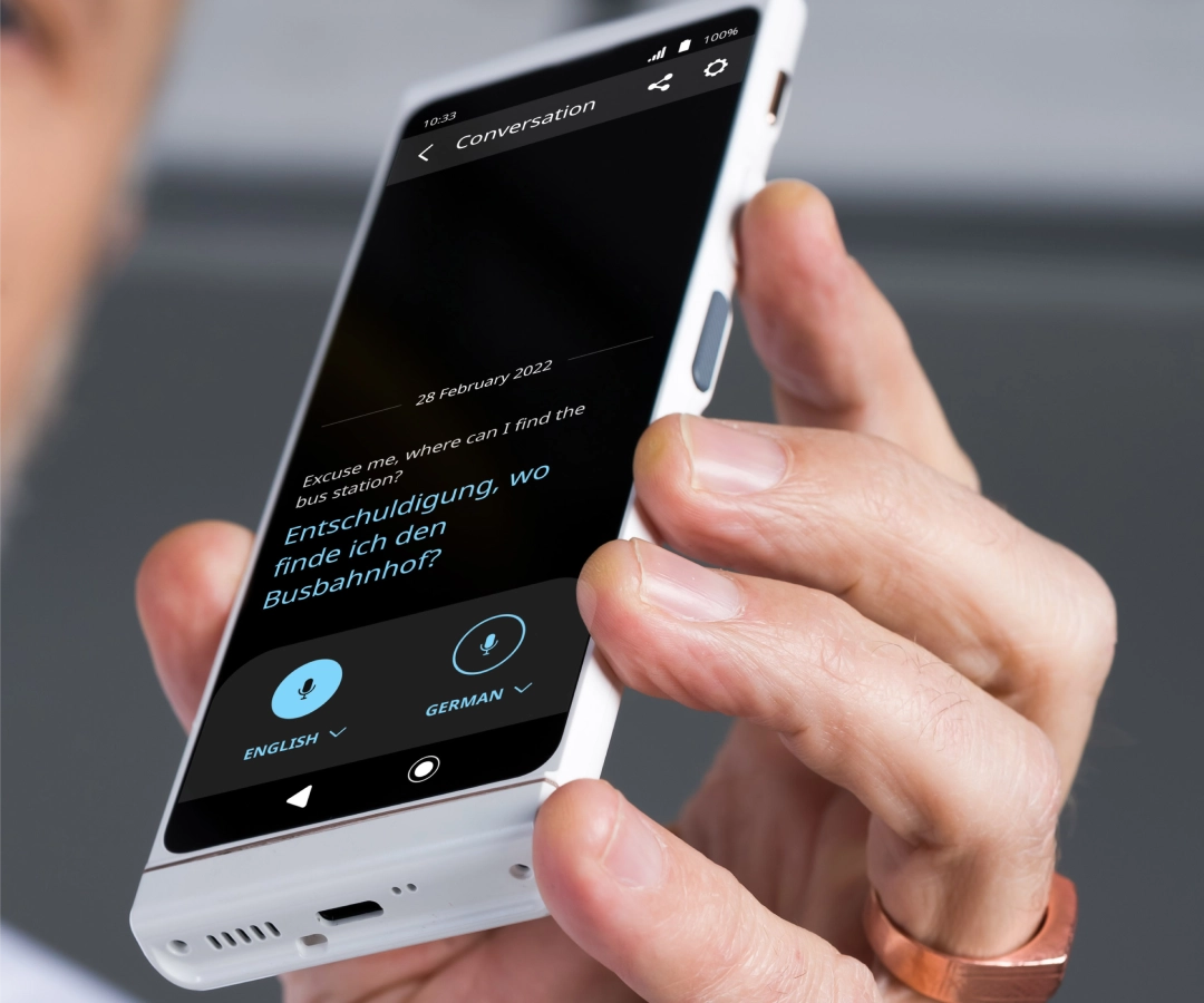 Vier Erwachsene, drei sichtbar und einer teilweise verdeckt, unterhalten sich im Freien am Wasser. Zwei Frauen, von denen eine lächelt und einen Hut trägt, zeigen der Gruppe etwas auf ihren Übersetzern, die sie in den Händen halten.