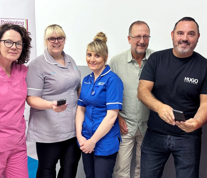 Fünf lächelnde Menschen stehen in Gesundheitskleidung nebeneinander, zwei von ihnen halten ein Übersetzergerät in der Hand.