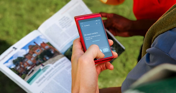 Eine Nahaufnahme einer Hand, die ein rotes elektronisches Gerät der Marke Vasco über einem aufgeschlagenen Reiseführer oder einer Broschüre hält. Auf dem Bildschirm des Geräts ist das Menü „Kontakte“ zu sehen, mit dem Text „Keine gespeicherten Kontakte. Fügen Sie jetzt oder nach einem Anruf einen neuen Kontakt hinzu.“ Es wird die Option „KONTAKT HINZUFÜGEN“ angezeigt.