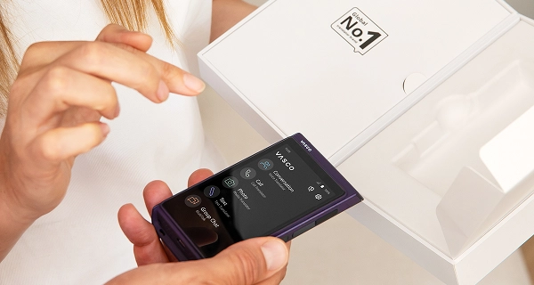 Eine Nahaufnahme von zwei Personen, die mit einem Gerät interagieren. Eine Person hält ein dunkelviolettes elektronisches Handgerät in der Hand, auf dessen Bildschirm eine numerische Wähltastatur zu sehen ist. Daneben steht eine Frau mit langen braunen Haaren und einem weißen Oberteil, die die Hand ausstreckt, um den Bildschirm zu berühren oder darauf zu zeigen. Im Hintergrund ist eine geöffnete weiße Produktverpackung zu sehen, auf deren Innenseite des Deckels ein grafisches Logo mit der Aufschrift „No. 1“ zu erkennen ist.