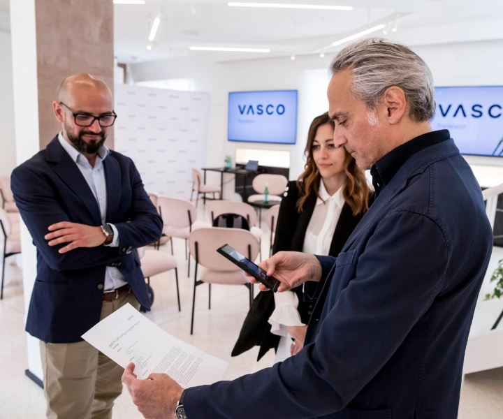 Drei professionell gekleidete Personen stehen in einem modernen Büro. Ein Mann hält ein Dokument in der Hand und benutzt seinen Übersetzer, während der andere Mann und eine Frau zuschauen. VASCO wird auf Bildschirmen im Hintergrund angezeigt.