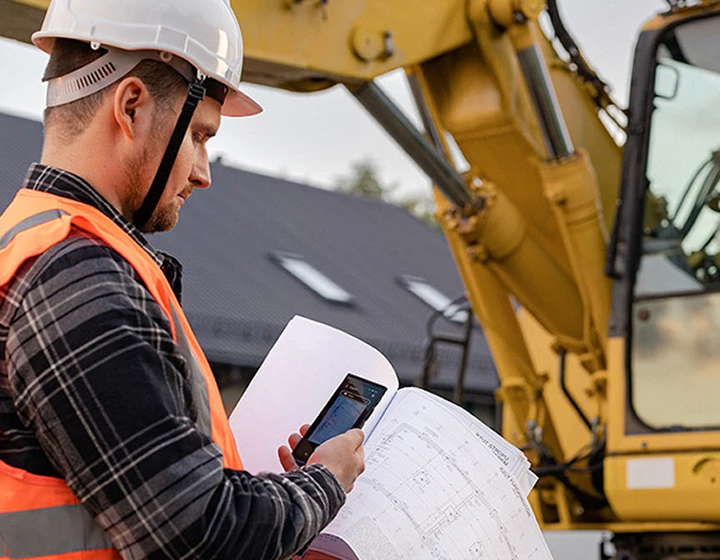 Ein Mann auf einer Baustelle in Helm und Arbeitskleidung fotografiert mit einem Vasco Translator V4 Baupläne. Im Hintergrund ist ein Bagger zu sehen.