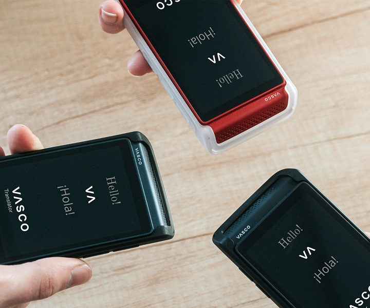 Drei Personen halten je einen Vasco Translator Q1 der Hand. Auf den Bildschirmen ist der Startbildschirm mit dem Vasco-Logo und den Worten „Hello!“ und „¡Hola!“ zu sehen. Die Geräte werden in Schutzhüllen in drei Farben gezeigt: 
