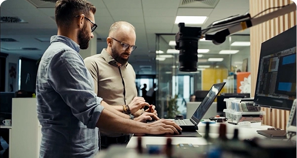 Zwei Männer mit Bart und Brille arbeiten gemeinsam an einem Stehpult in einem modernen Büro oder Labor. Der eine Mann in einem hellblauen Hemd bedient einen Laptop, während der andere Mann in einem beigen Hemd neben ihm steht und auf ein kleines elektronisches Bauteil in seiner Hand zeigt. Das Pult ist mit einem großen Monitor, verschiedenen elektronischen Geräten und einer an einem verstellbaren Arm befestigten Kamera ausgestattet. Im Hintergrund ist ein heller, offener Arbeitsbereich mit Glaswänden zu sehen. 