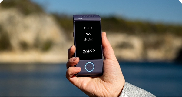 Eine Nahaufnahme einer Hand, die ein violettes elektronisches Übersetzungsgerät für Baskisch vor einem hellen Außenhintergrund hält. Auf dem Bildschirm sind die Begrüßungen „Hello!“, „¡Hola!“ und das Logo „VASCO Translator“ zu sehen. Im Hintergrund ist eine sonnenbeschienene Küstenlandschaft mit blauem Wasser und hellen Felsklippen unter einem klaren Himmel in Weichzeichneroptik zu sehen. Die Person, die das Gerät hält, trägt einen hellgrauen oder silberfarbenen, strukturierten Jackenärmel. 