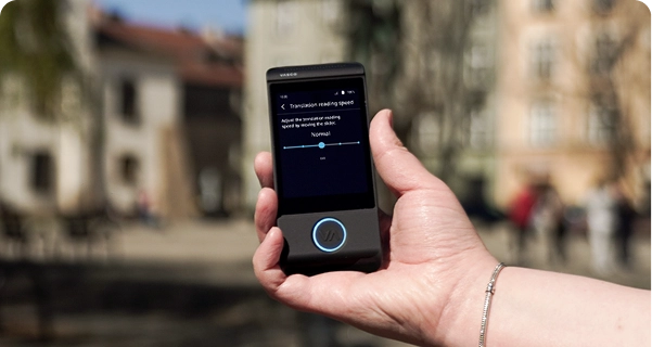 Nahaufnahme der Hand einer Person, die einen schwarzen elektronischen Übersetzer der Marke Vasco hält, vor dem verschwommenen Hintergrund eines Stadtplatzes mit historischen Gebäuden. Auf dem Bildschirm des Geräts sind die Einstellungen für die „Lesegeschwindigkeit der Übersetzung“ zu sehen, darunter ein Schieberegler, der auf „Normal“ eingestellt ist, sowie der Hinweis: „Passen Sie die Lesegeschwindigkeit der Übersetzung an, indem Sie den Schieberegler verschieben.“ Die Person, die das Gerät hält, trägt ein silbernes Armband. Das helle Tageslicht hebt die Benutzeroberfläche des Bildschirms hervor, sodass die Einstellungen gut zu erkennen sind. 