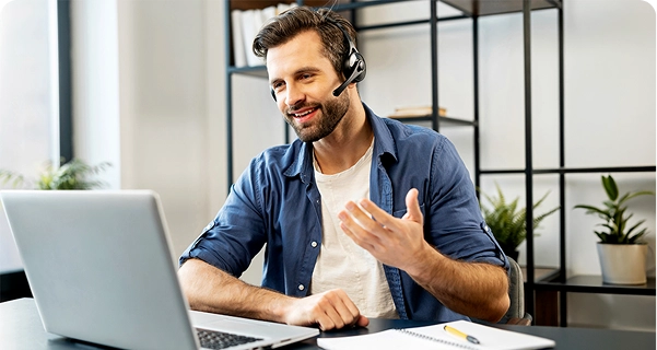 Ein freundlich aussehender Mann mit Bart und dunklem Haar sitzt an einem Schreibtisch und trägt ein professionelles Headset mit Mikrofon. Er lächelt und gestikuliert mit einer Hand, während er auf den Bildschirm eines Laptops schaut; es sieht so aus, als befände er sich mitten in einem Videoanruf oder leiste technischen Support. Er trägt ein blaues Hemd über einem weißen T-Shirt. Auf dem Schreibtisch vor ihm liegen ein Laptop, ein Notizbuch und ein gelber Stift. Der Hintergrund ist ein heller, moderner Büroraum mit einem minimalistischen schwarzen Metallregal und grünen Topfpflanzen. 