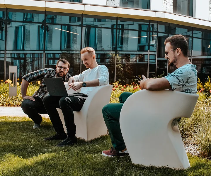 Drei Männer sitzen auf modernen weißen Stühlen vor einem Gebäude und arbeiten gemeinsam an einem Laptop. Sie wirken entspannt und konzentriert; einer von ihnen lächelt, während er seinen Übersetzer ansieht. Sie sind von üppigem Gras und Blumen umgeben.