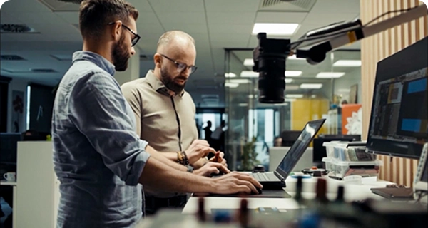 Zwei Männer mit Bärten und Brillen arbeiten gemeinsam an einem Stehpult in einem modernen Büro oder Labor. Der eine, in einem hellblauen Hemd, benutzt einen Laptop, während der andere, in einem beigen Hemd, neben ihm steht und auf ein kleines elektronisches Bauteil in seiner Hand deutet. Das Pult ist mit einem großen Monitor, verschiedenen elektronischen Geräten und einer an einem verstellbaren Arm befestigten Kamera ausgestattet. Im Hintergrund sieht man einen hellen, offenen Arbeitsbereich mit Glaswänden.