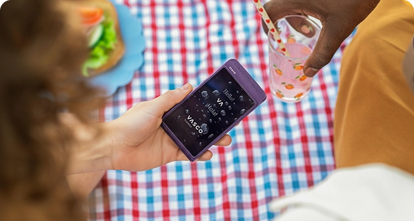 Eine Draufsicht auf einen violetten Vasco Translator Q1, der über einer Picknickdecke gehalten wird. Der Bildschirm des Geräts ist sichtbar nass und mit Wassertropfen bedeckt, was seine Wasserbeständigkeit für den Einsatz im Freien beweist. Im Hintergrund sind ein Sandwich und eine Person mit einem Glas zu sehen.