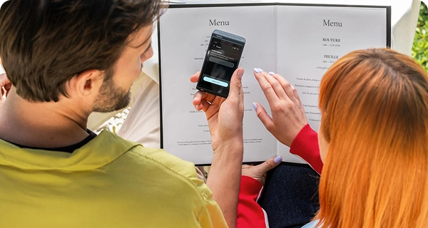Eine Kameraaufnahme aus der Schulterperspektive zeigt einen Mann und eine Frau mit leuchtend orangefarbenen Haaren, die an einem Restauranttisch sitzen und die Speisekarte studieren. Der Mann hält einen schwarzen Vasco Translator Q1 in der Hand, auf dem ein Bildschirm mit der Funktion „Teilen“ und einem Eingabefeld zum Versenden der Übersetzung per E-Mail angezeigt wird. Im Hintergrund sind eine weiße Tischdecke und das übrige Restaurantgeschirr zu sehen.